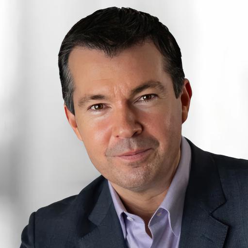 A man in a dark suit and light purple shirt is posing in front of a plain white background.