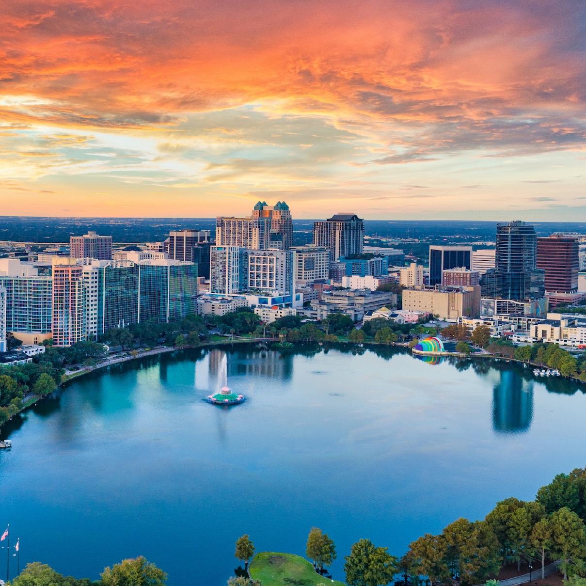 Aerial view of a city skyline at sunset with a large lake and surrounding trees in the foreground.