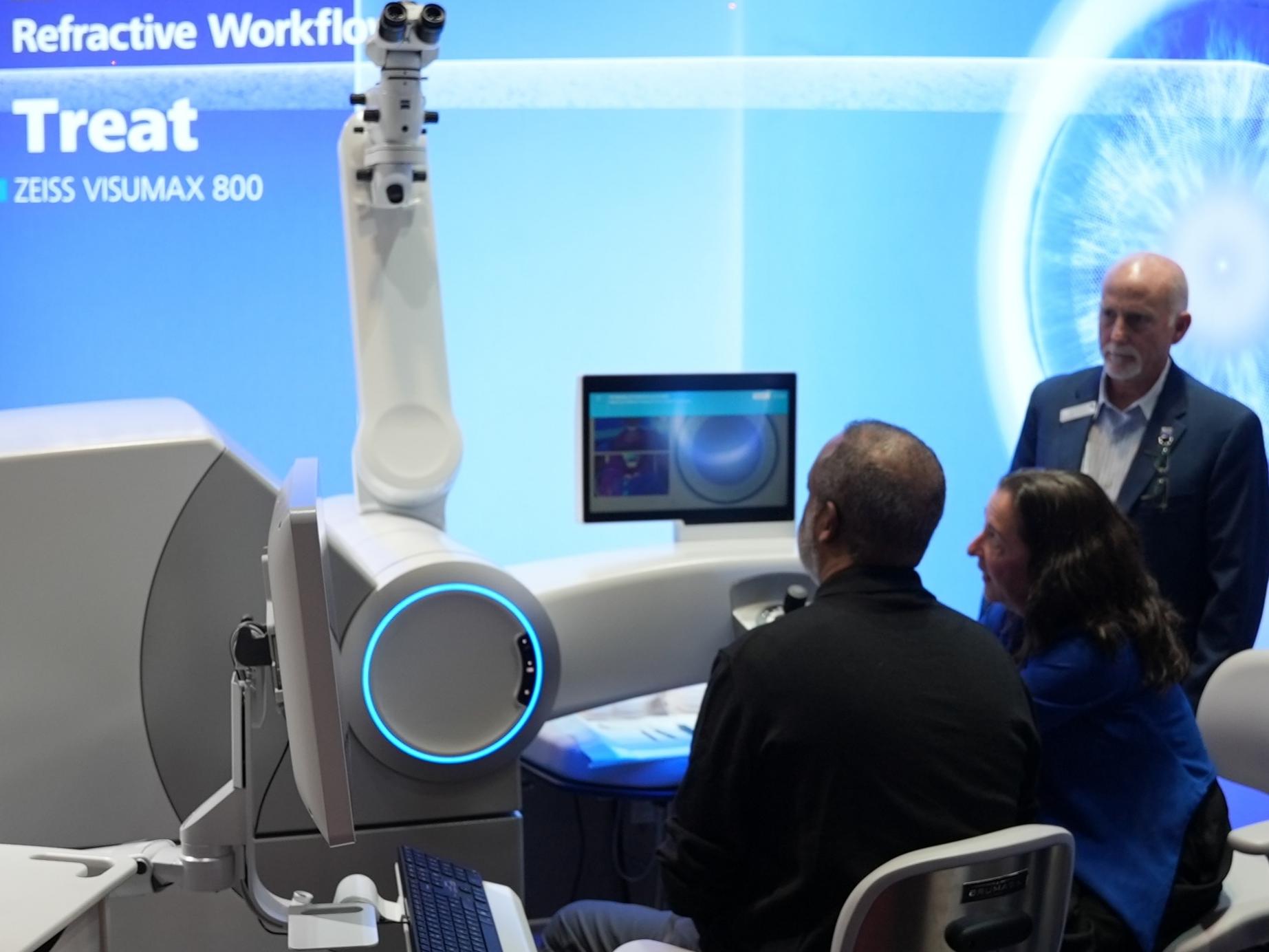 A group of people is gathered around the ZEISS VISUMAX 800 laser system in a clinical setting with a large display showcasing the ZEISS Corneal Refractive Workflow.