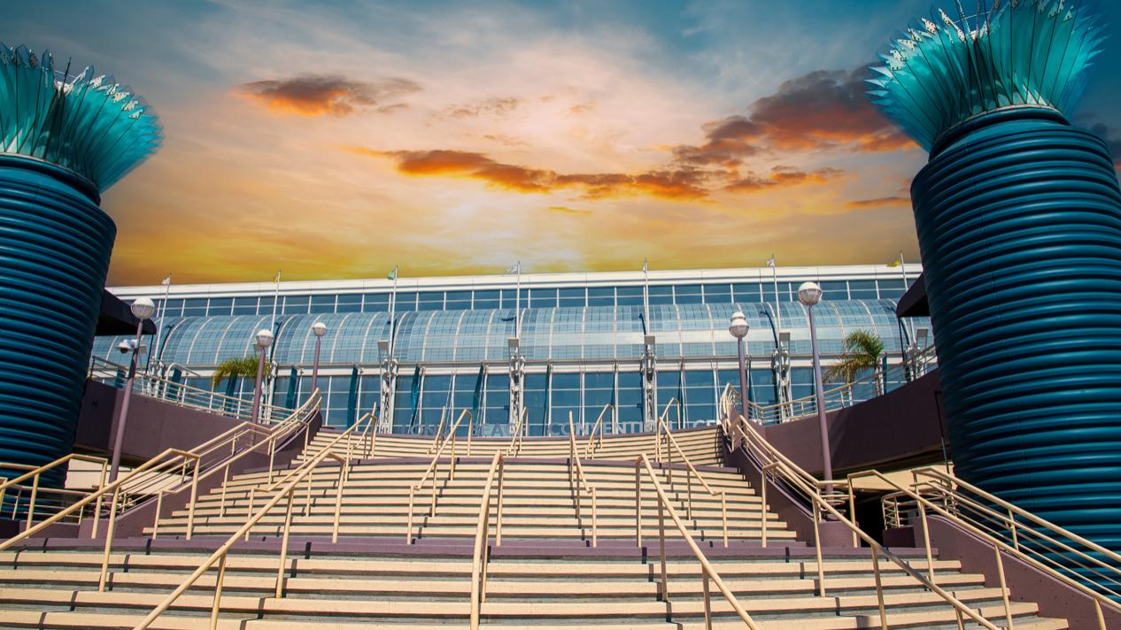 Wide staircase leading up to a modern glass building with two large blue cylindrical structures on either side, set against a dramatic sunset sky.