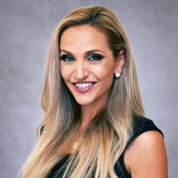 Professional woman with long blonde hair wearing a sleeveless black dress, smiling in front of a neutral gray background.