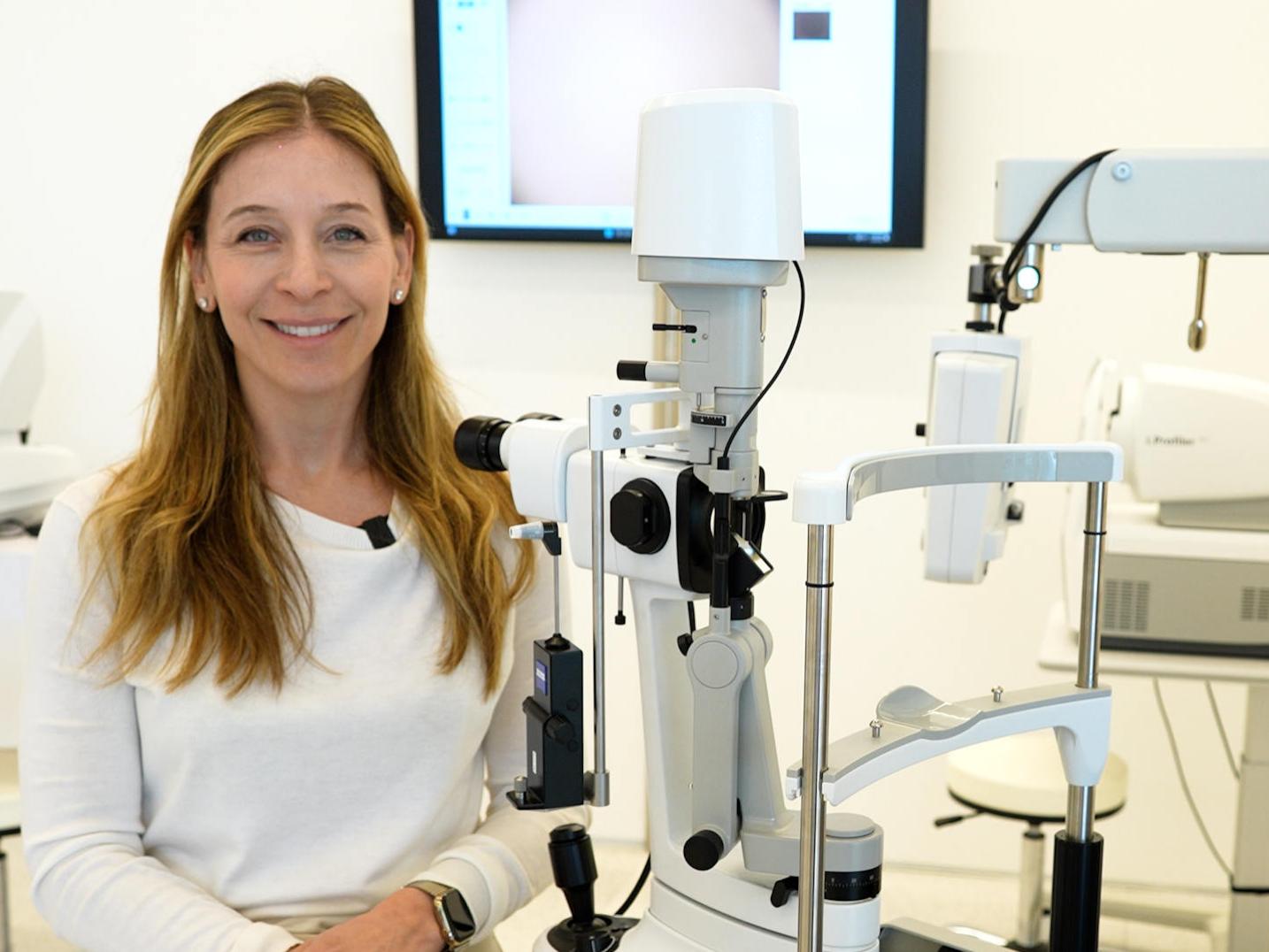 A woman is smiling while seated next to a ZEISS ophthalmic diagnostic device in a modern clinical setting.