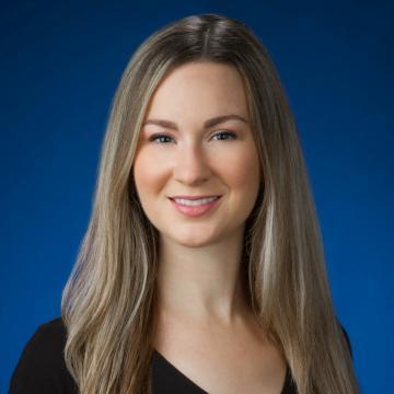 Professional headshot of a woman with long blonde hair wearing a black top, in front of a blue background, for ZEISS.