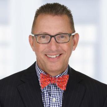 Smiling man wearing glasses, a dark pinstripe suit, a blue checkered shirt, and a red bow tie, in front of a blurred office background.