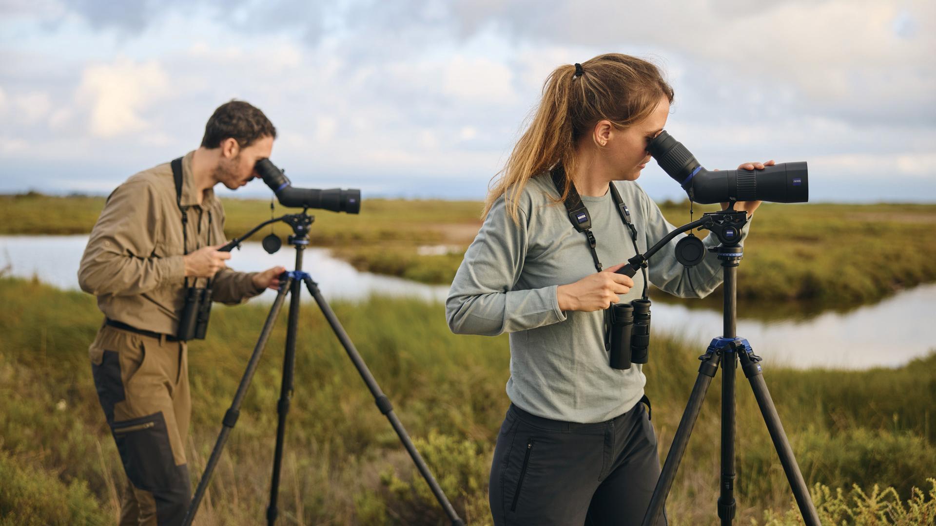 Two people outdoors using the new ZEISS Conquest Apia spotting scopes mounted on tripods to observe the open landscape with grass, low vegetation, and a small body of water in the background.