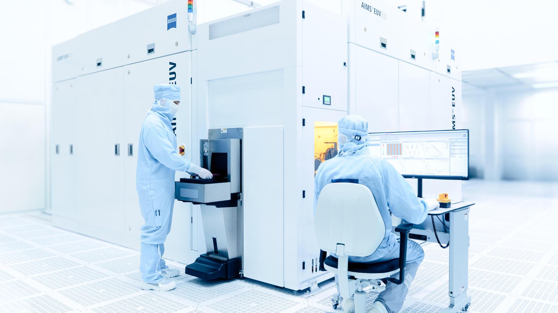 In a white, brightly lit cleanroom stands a device for mask qualification. Two people in protective suits are working on the device.