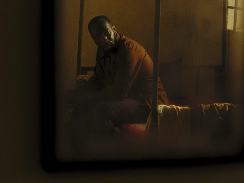 A man wearing a gray blazer, black shirt, chain necklace, and ripped jeans sits in a modern, dimly lit room with large windows and indoor plants, captured with ZEISS lens clarity.