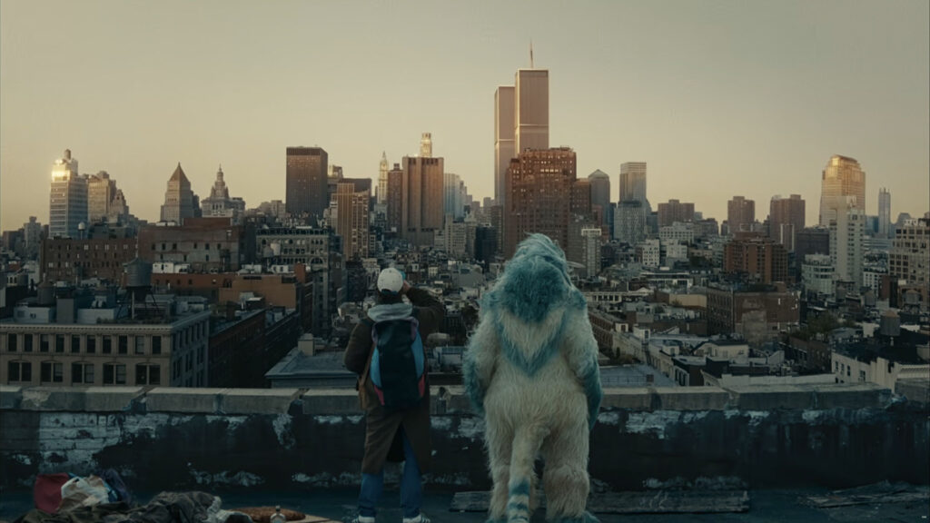 A man wearing a gray blazer, black shirt, chain necklace, and ripped jeans sits in a modern, dimly lit room with large windows and indoor plants, captured with ZEISS lens clarity.