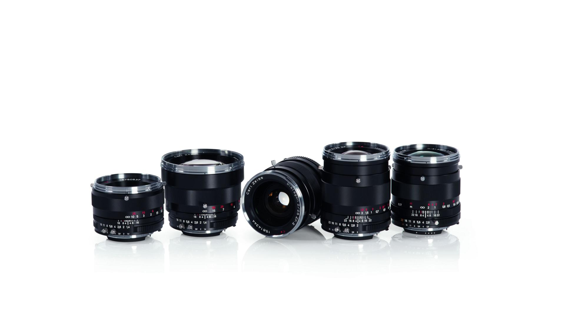 Five black ZEISS camera lenses are arranged in a row on a reflective white surface.