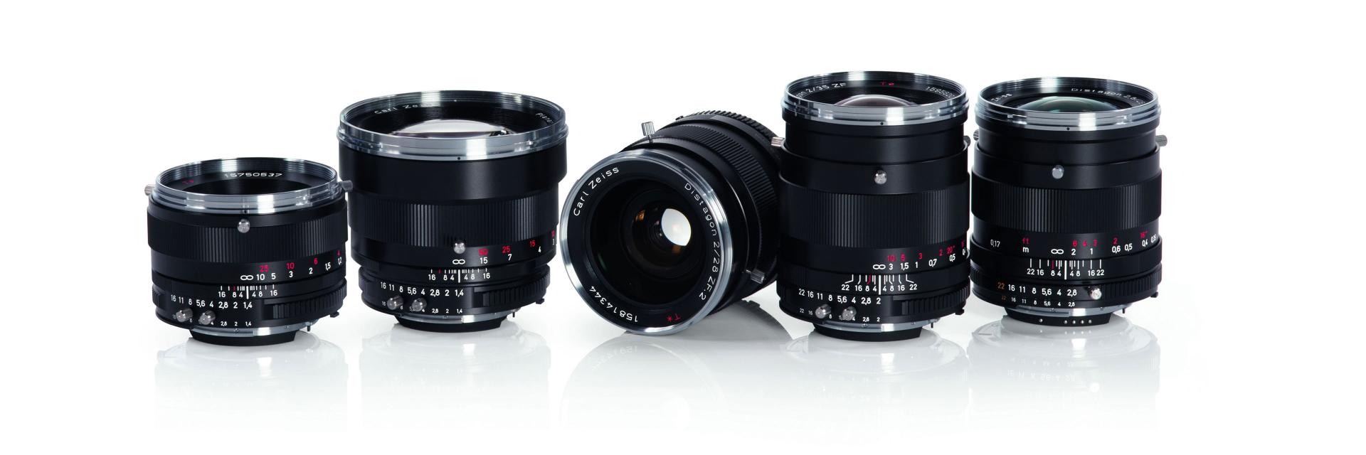 Five black ZEISS camera lenses are arranged in a row on a reflective white surface.