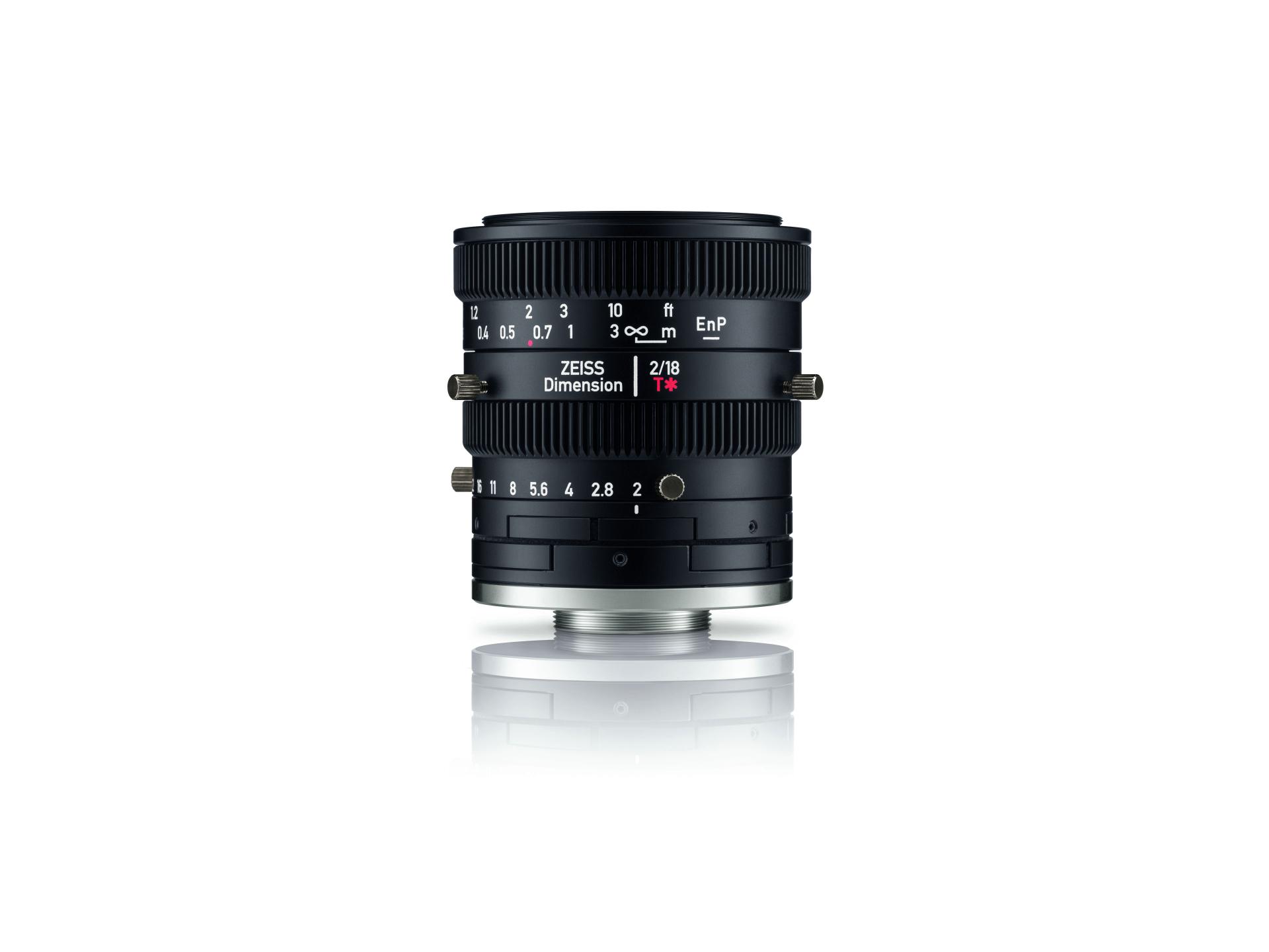 Black ZEISS Dimension 2/18 E-mount camera lens with marked focus and aperture adjustment rings, shown standing upright on a white reflective surface.