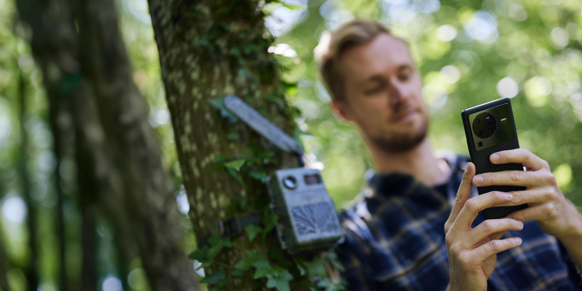 Un uomo è seduto su un divano di pelle e usa uno smartphone per guardare immagini di animali selvatici; sullo sfondo si vedono un tavolino da caffè in legno e un tappeto di pelle di mucca.