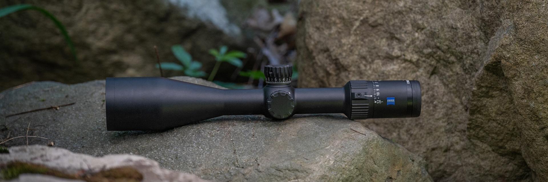A ZEISS riflescope is displayed lying on a rock outdoors, surrounded by natural elements like stones and plants.