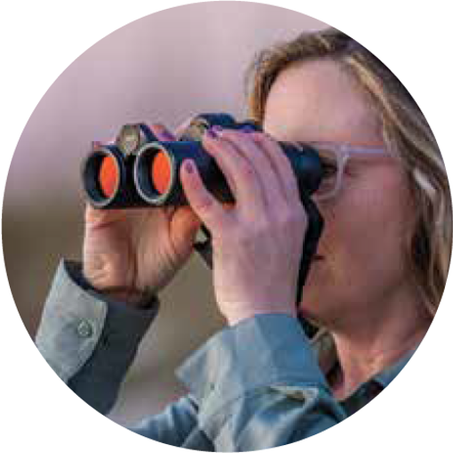 A woman wearing glasses looks through ZEISS binoculars with orange-tinted lenses.