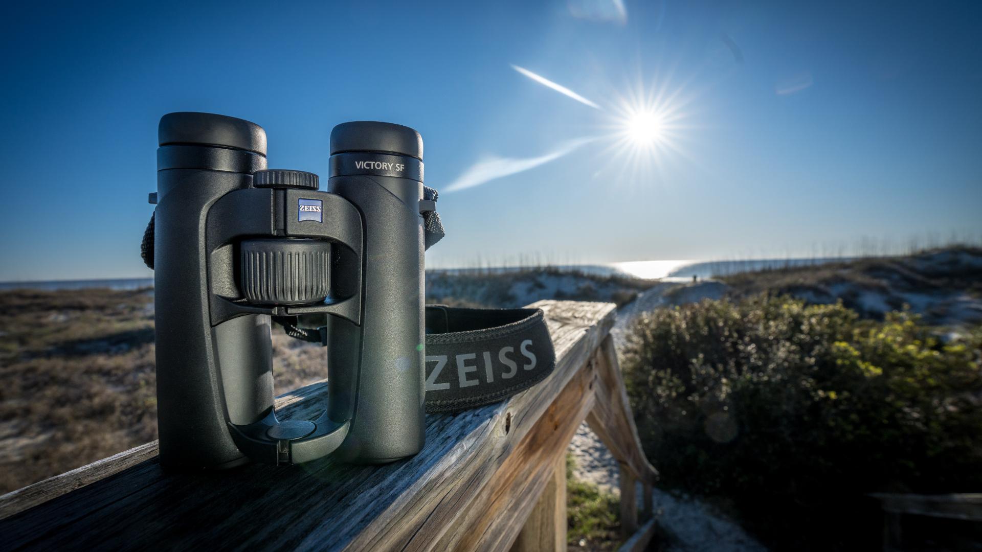 A pair of ZEISS binoculars resting on a wooden railing with a coastal landscape and bright sun in the background.