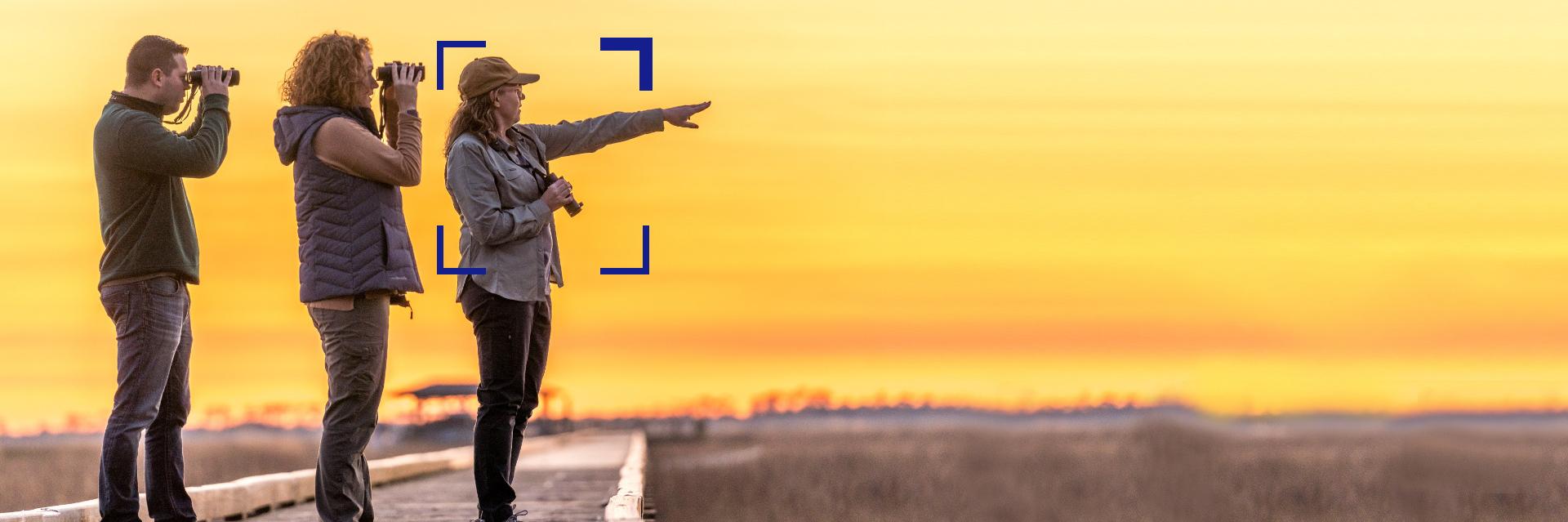 Three people stand on a wooden pathway at sunset, two using binoculars and one pointing into the distance, with a ZEISS focus frame highlighting the center figure.