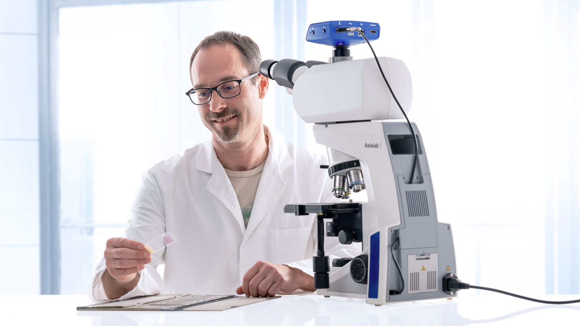 Scientist in lab coat examining a microscope slide infront of the Axiolab 5 microscope