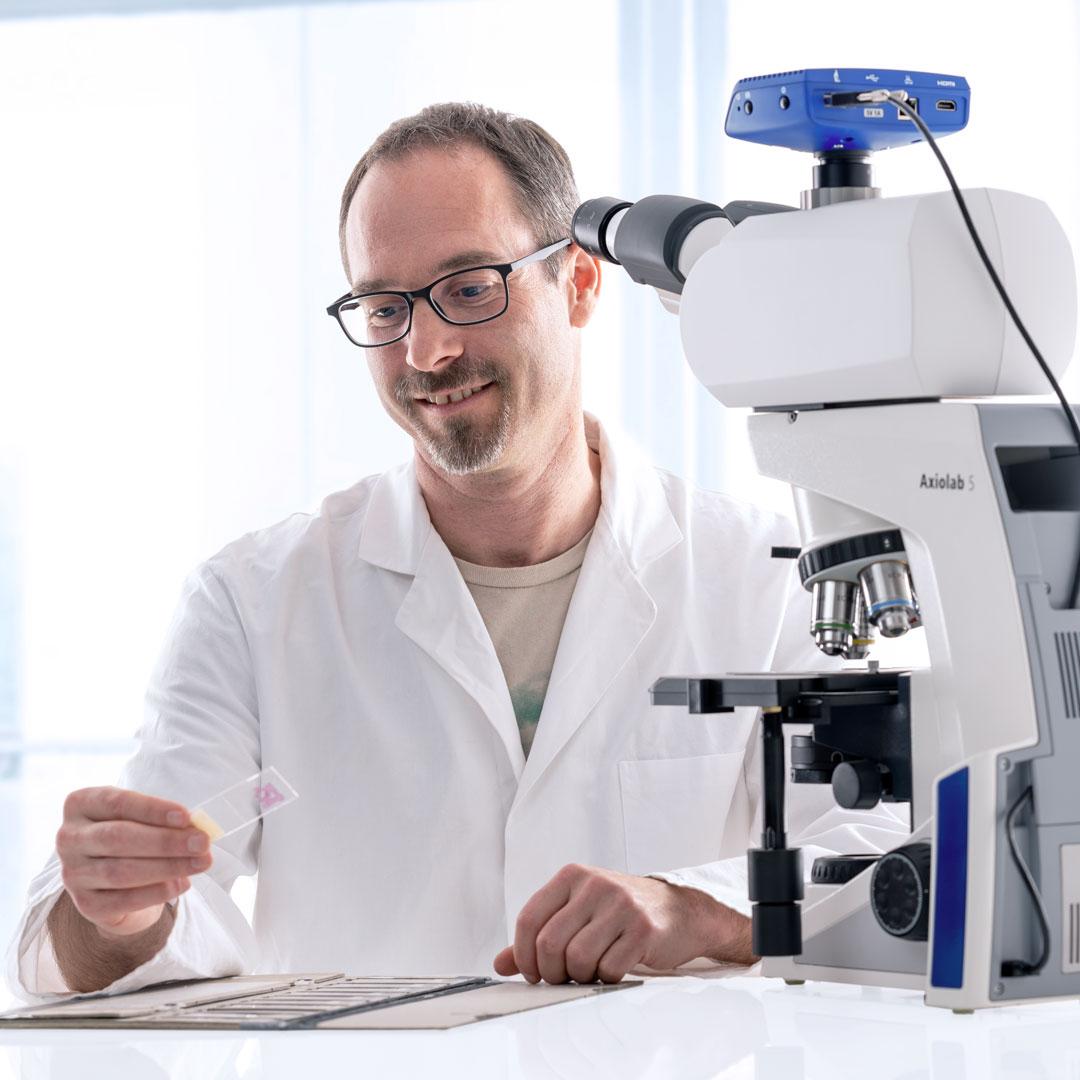 Scientist in lab coat examining a microscope slide infront of the Axiolab 5 microscope