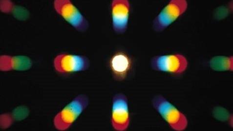 A central white light surrounded by eight multicolored, elongated light blurs and further light blurs on the very left and right, forming a radial pattern on a black background. 