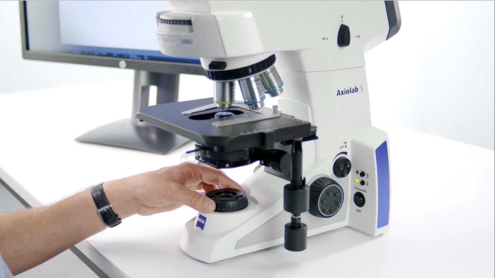 Image of a ZEISS Axiolab 5 microscope on a white table with a male person's arm in the foreground adjusting the field stop diaphragm. A screen can be seen in the background.
