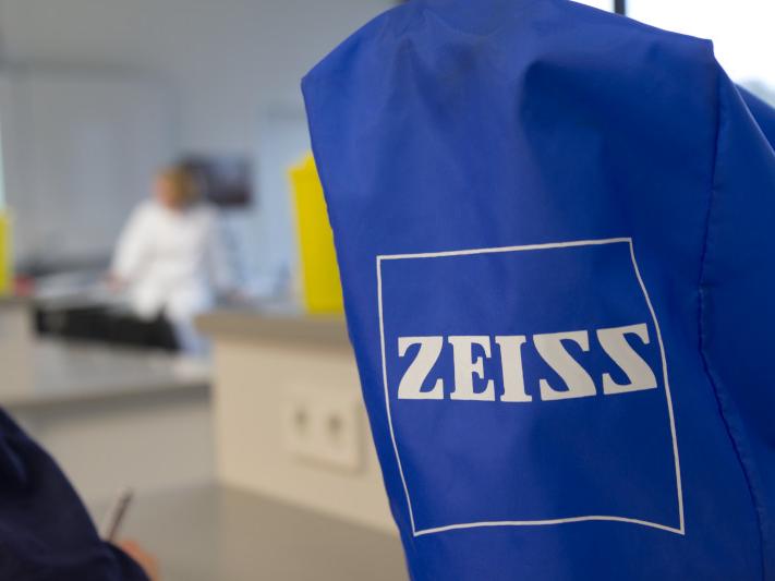A blue ZEISS equipment cover is in focus in the foreground, with a blurred laboratory setting and a person in a white coat in the background.