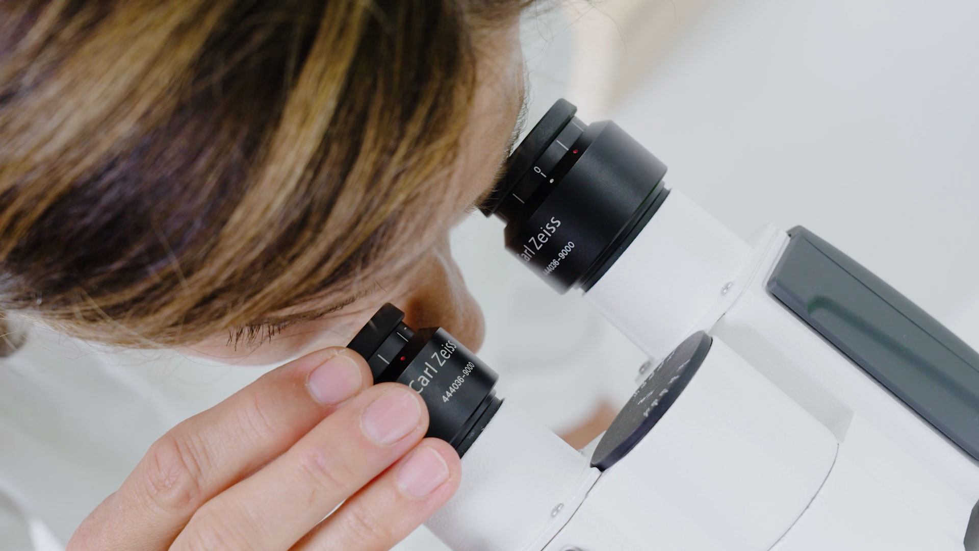 Woman in a lab adjusting a microscope while looking through it.