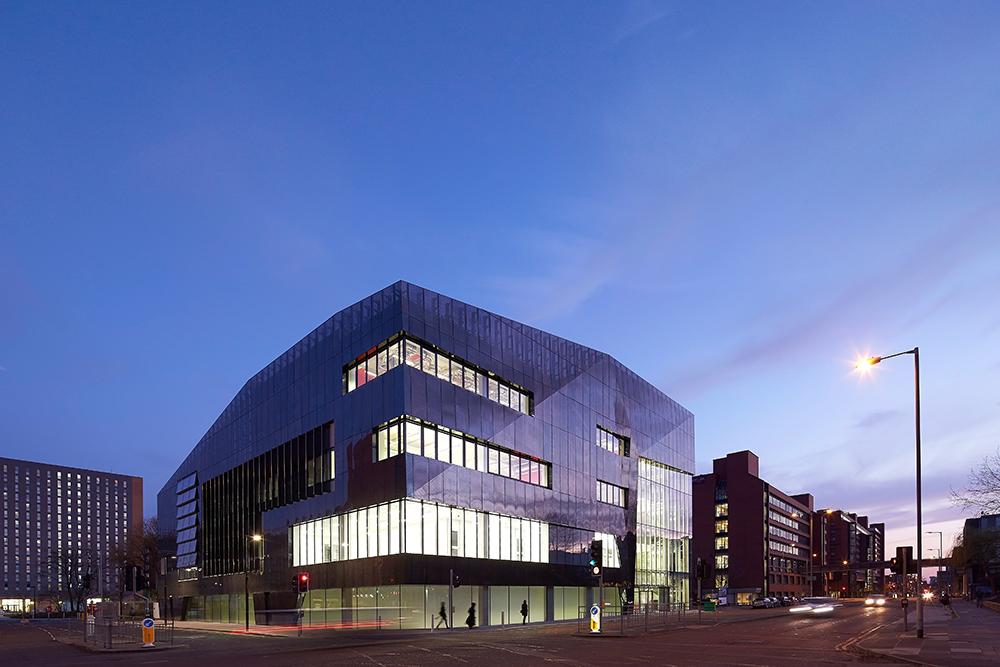 A modern glass building at a street corner is illuminated at dusk, with adjacent buildings and streetlights visible under a clear evening sky.