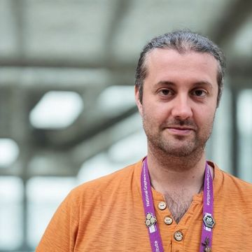 A man with long hair tied back, wearing an orange shirt and a purple lanyard, stands indoors with a blurred background.