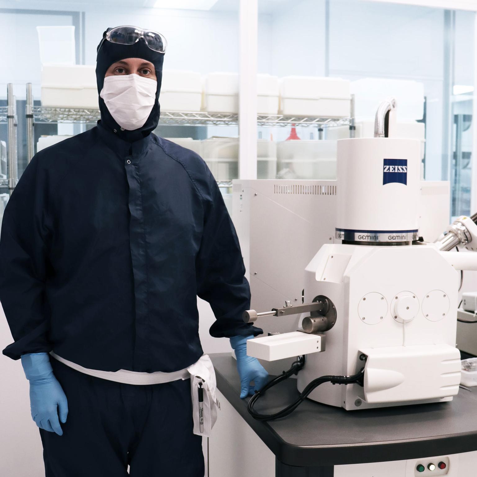 A person in cleanroom attire stands next to a ZEISS Sigma scanning electron microscope in a laboratory setting.