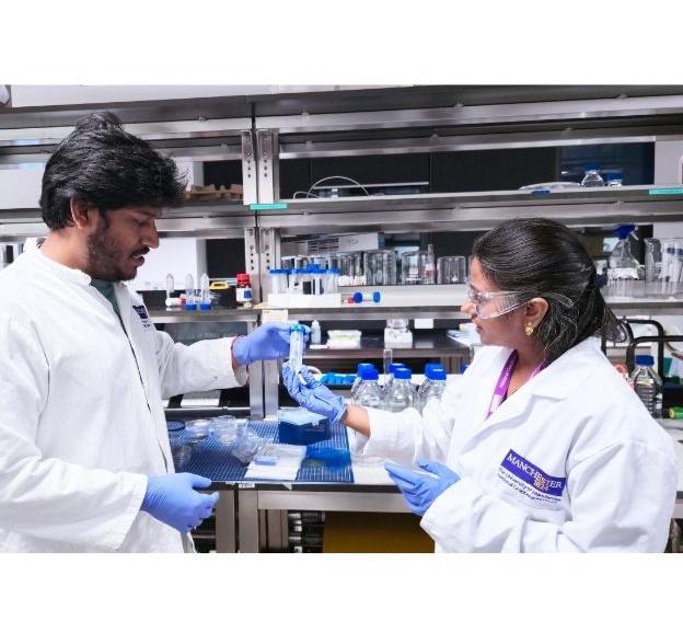 Two scientists wearing lab coats and gloves work together in a laboratory, handling a test tube surrounded by various lab equipment on shelves.