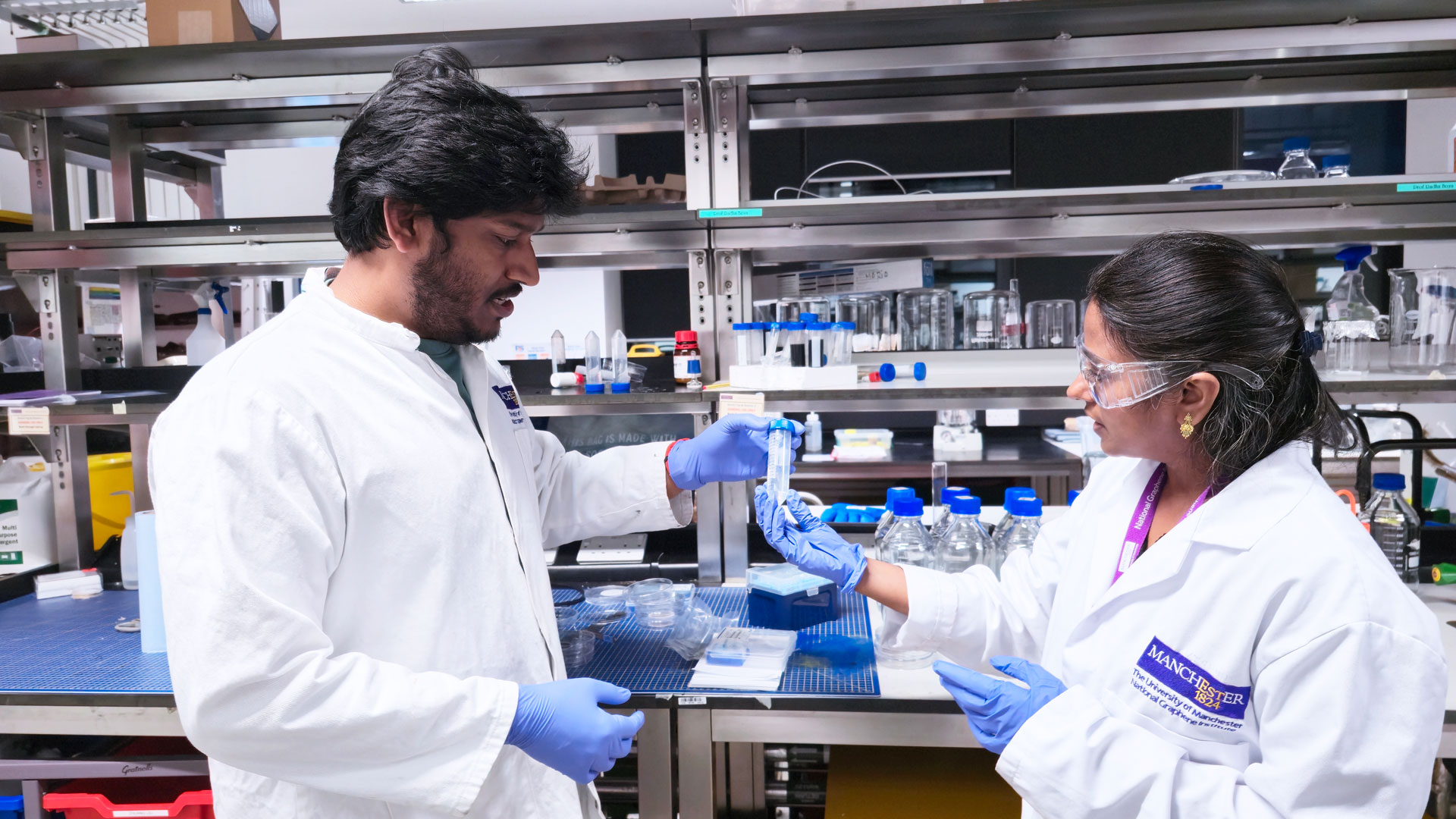 Two scientists wearing lab coats and gloves work together in a laboratory, handling a test tube surrounded by various lab equipment on shelves.