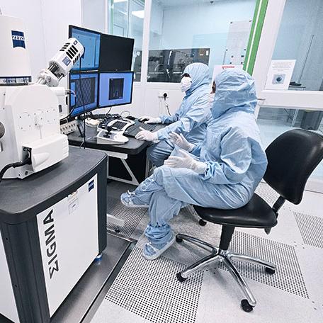 Two individuals in blue cleanroom suits operate a ZEISS Sigma microscope at a workstation with multiple monitors inside a laboratory environment.