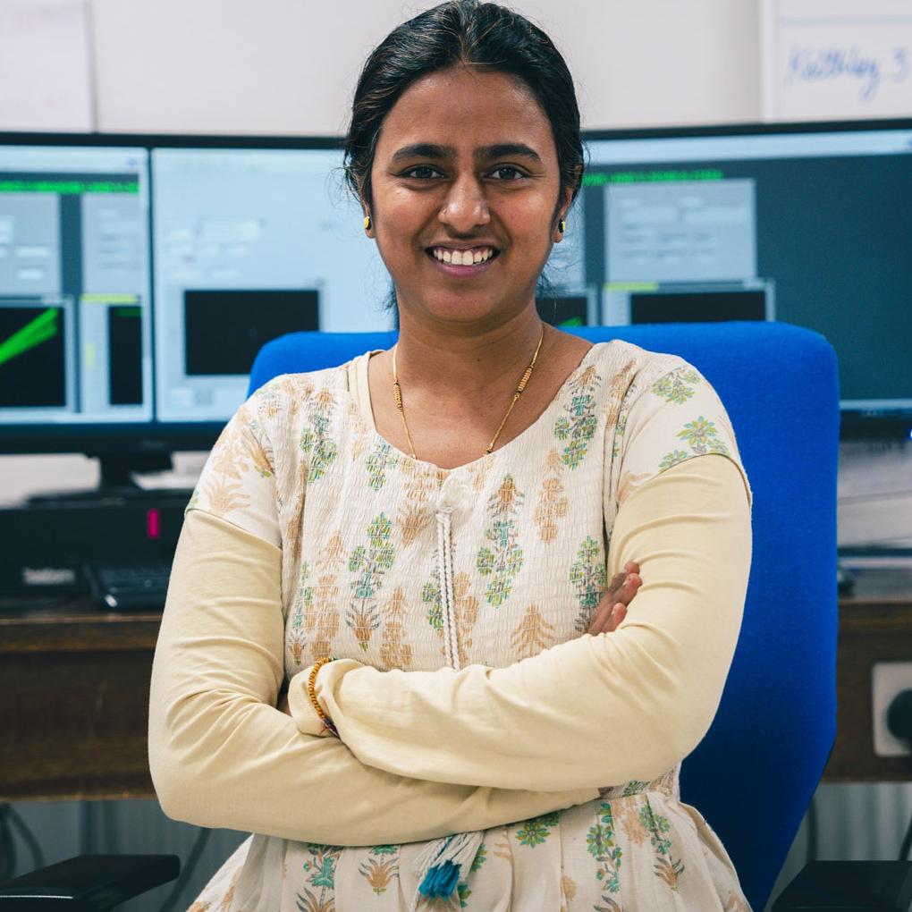 A woman sits with arms crossed, smiling in an office with computer monitors, equipment, and a whiteboard in the background.