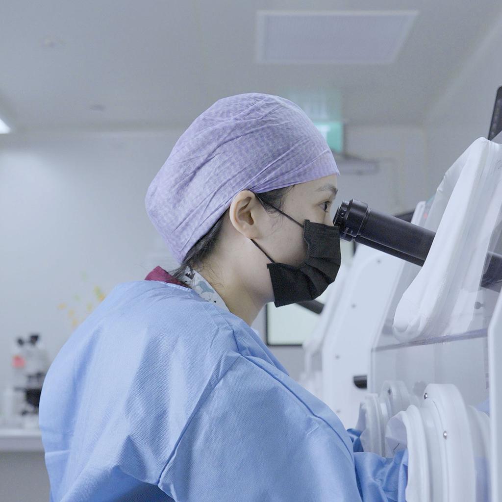 Person in a blue gown and purple cap uses a microscope in a laboratory setting.