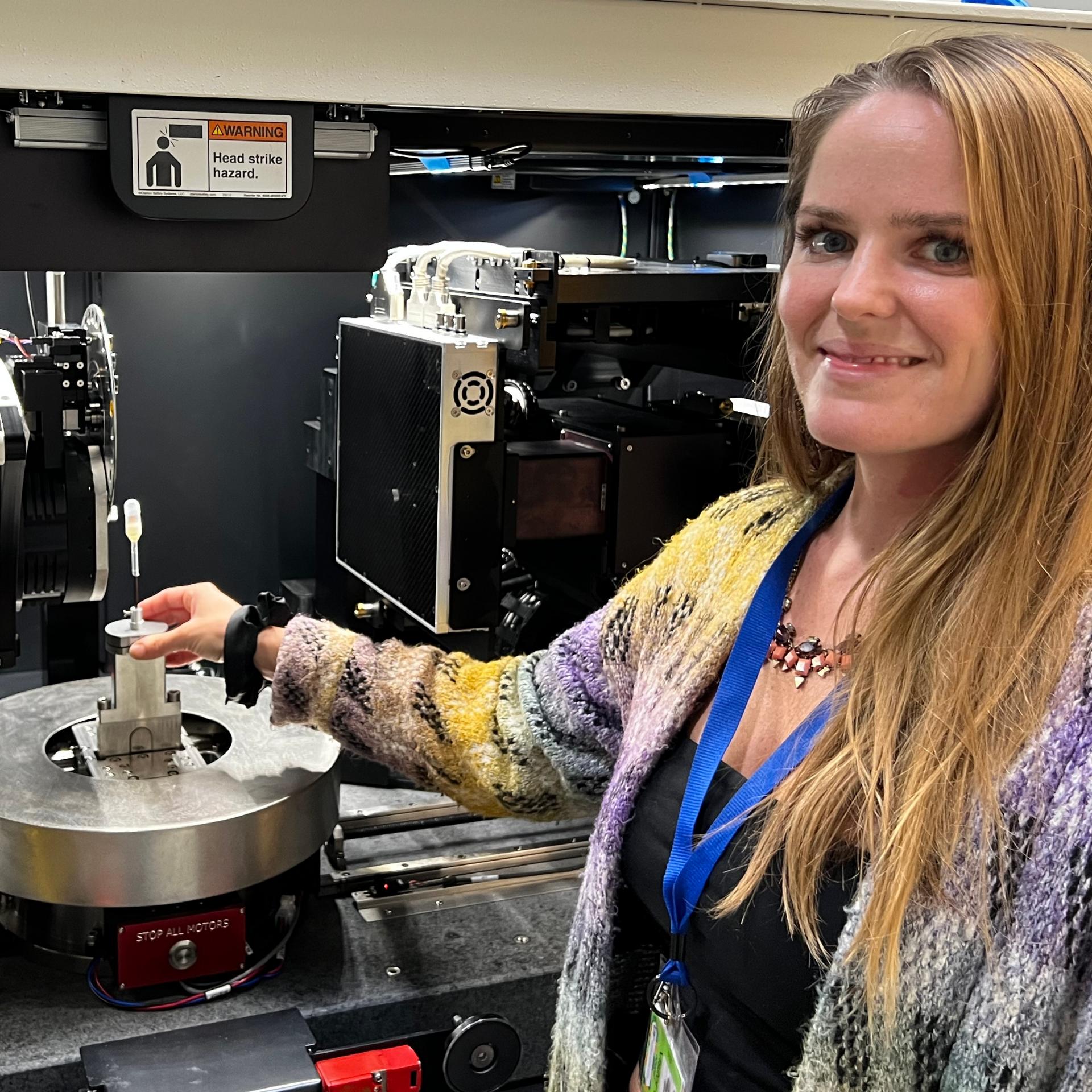 A smiling woman stands beside a X-Ray microscope ZEISS Xradia Versa 620, holding a sample holder. She is wearing a colorful sweater and a blue lanyard.