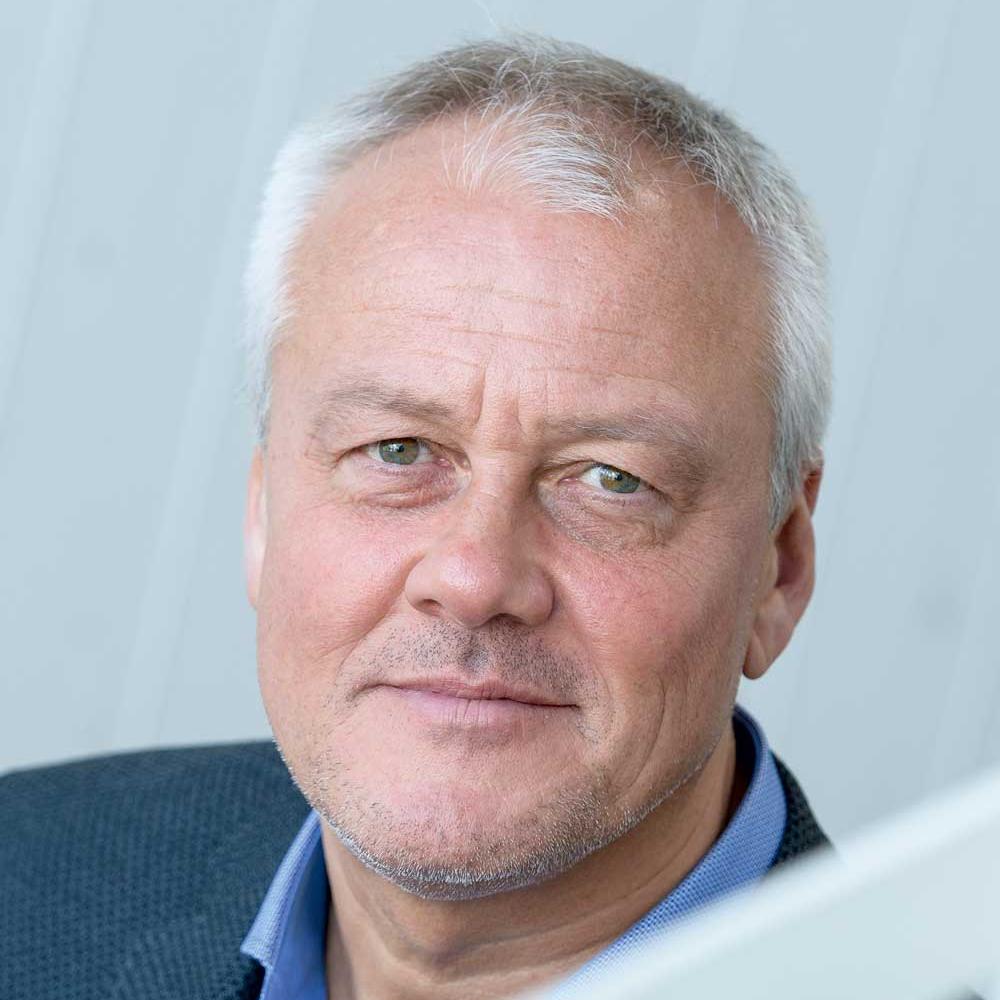 A middle-aged man with short gray hair and light stubble, wearing a blue shirt and dark jacket, looks at the camera with a neutral expression against a pale background.