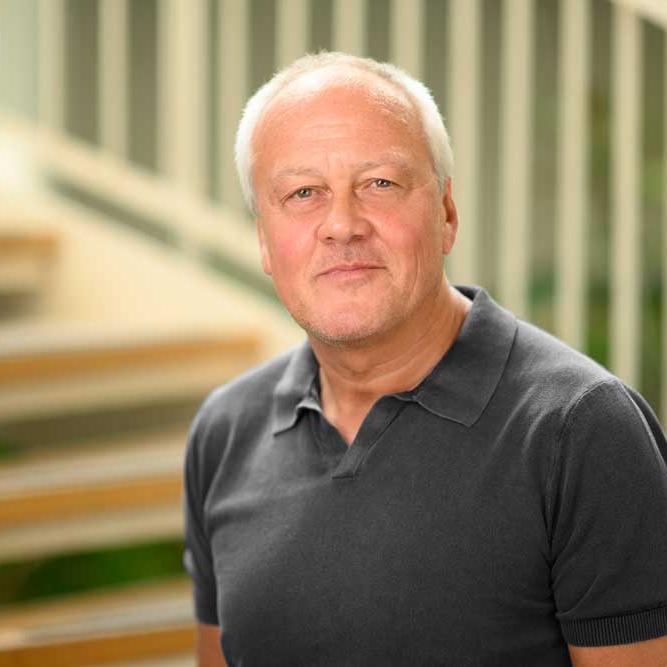 A middle-aged man with short gray hair wearing a dark polo shirt stands in front of a staircase with a curved railing.