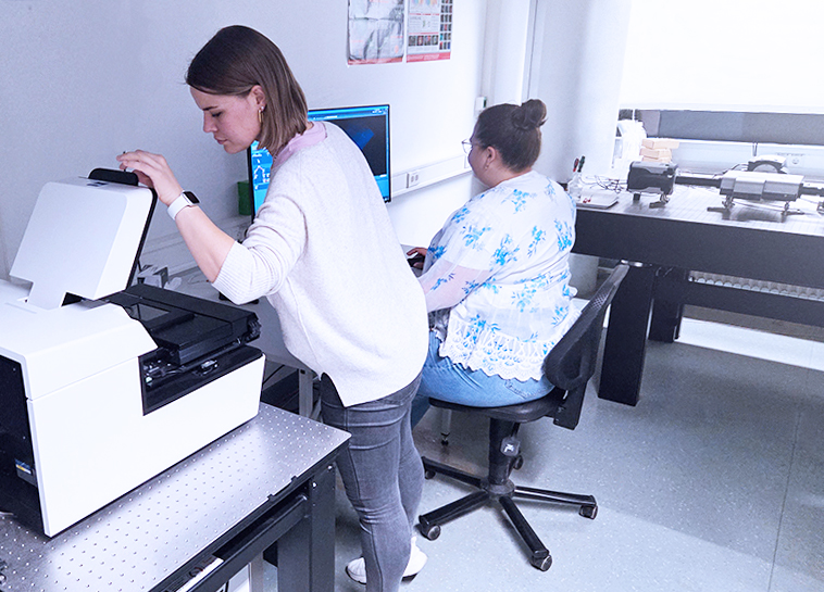 Two women work in a lab; one operates a large machine on a table, while the other sits at a computer desk with multiple monitors.