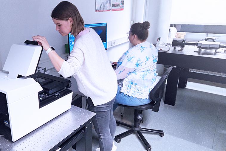 Two women work in a lab; one operates a large machine on a table, while the other sits at a computer desk with multiple monitors.