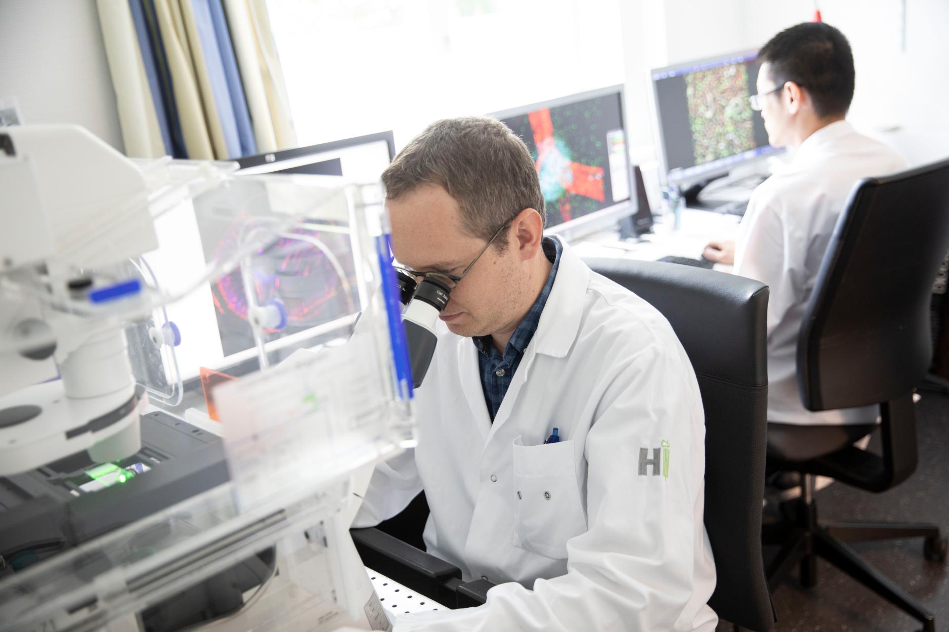 Two scientists in lab coats work in a lab; one looks into a microscope, the other works at a computer with screens displaying colorful images.