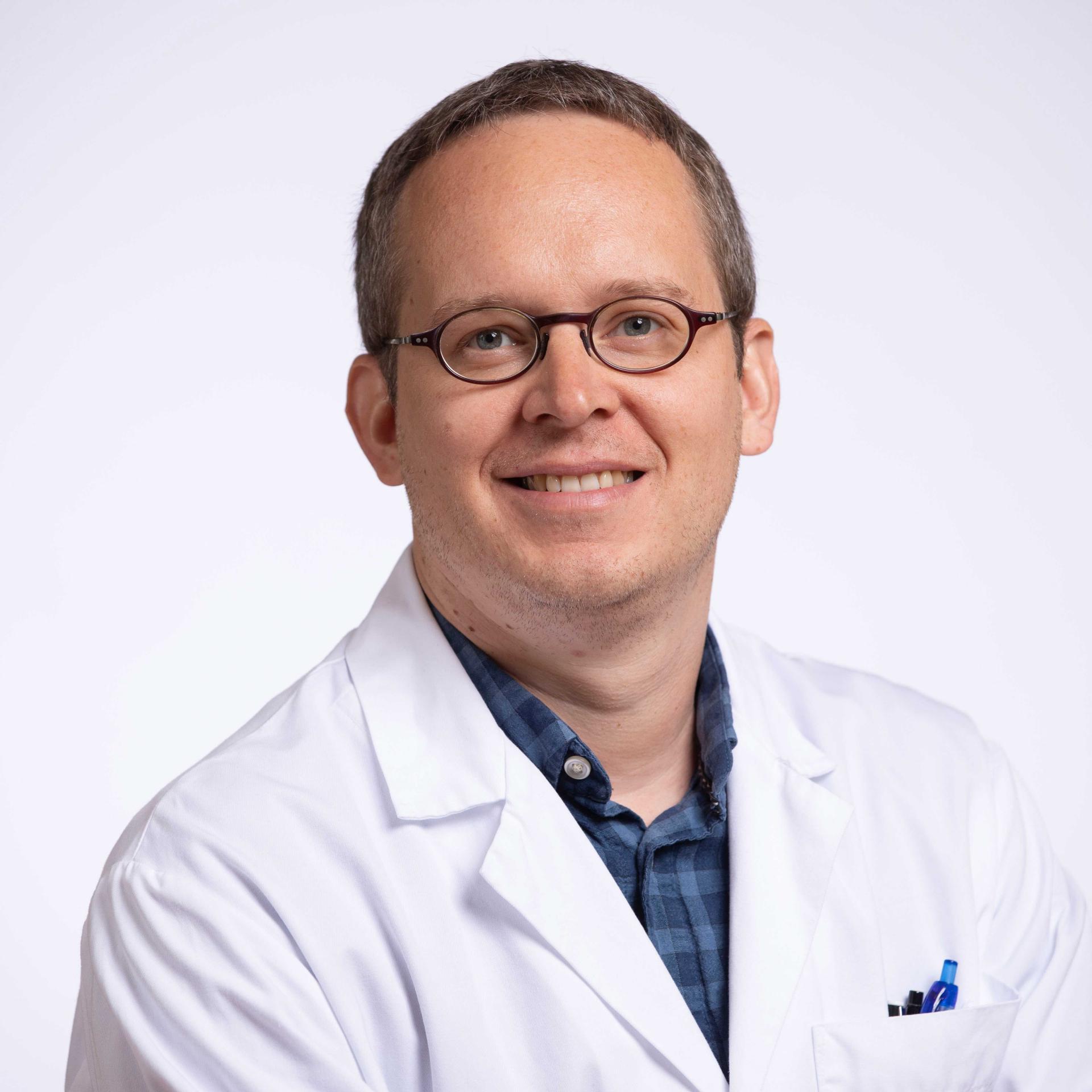 Man with glasses in a white lab coat and shirt stands in front of a light background