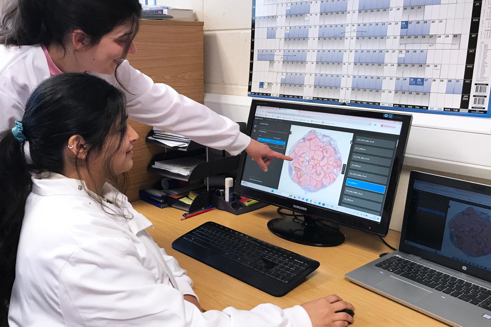 Two lab technicians in white coats examine a digital image of a cross-section on a computer screen. One technician is seated while the other points to the screen.