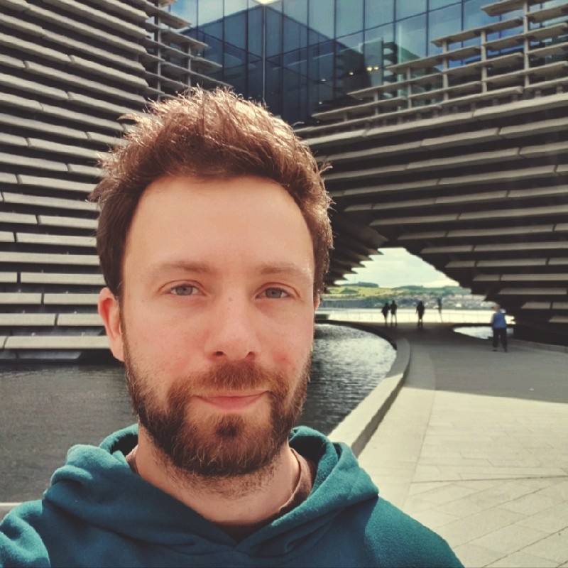 A person with a beard and a hoodie stands in front of a modern, angular building with reflective glass and water features.