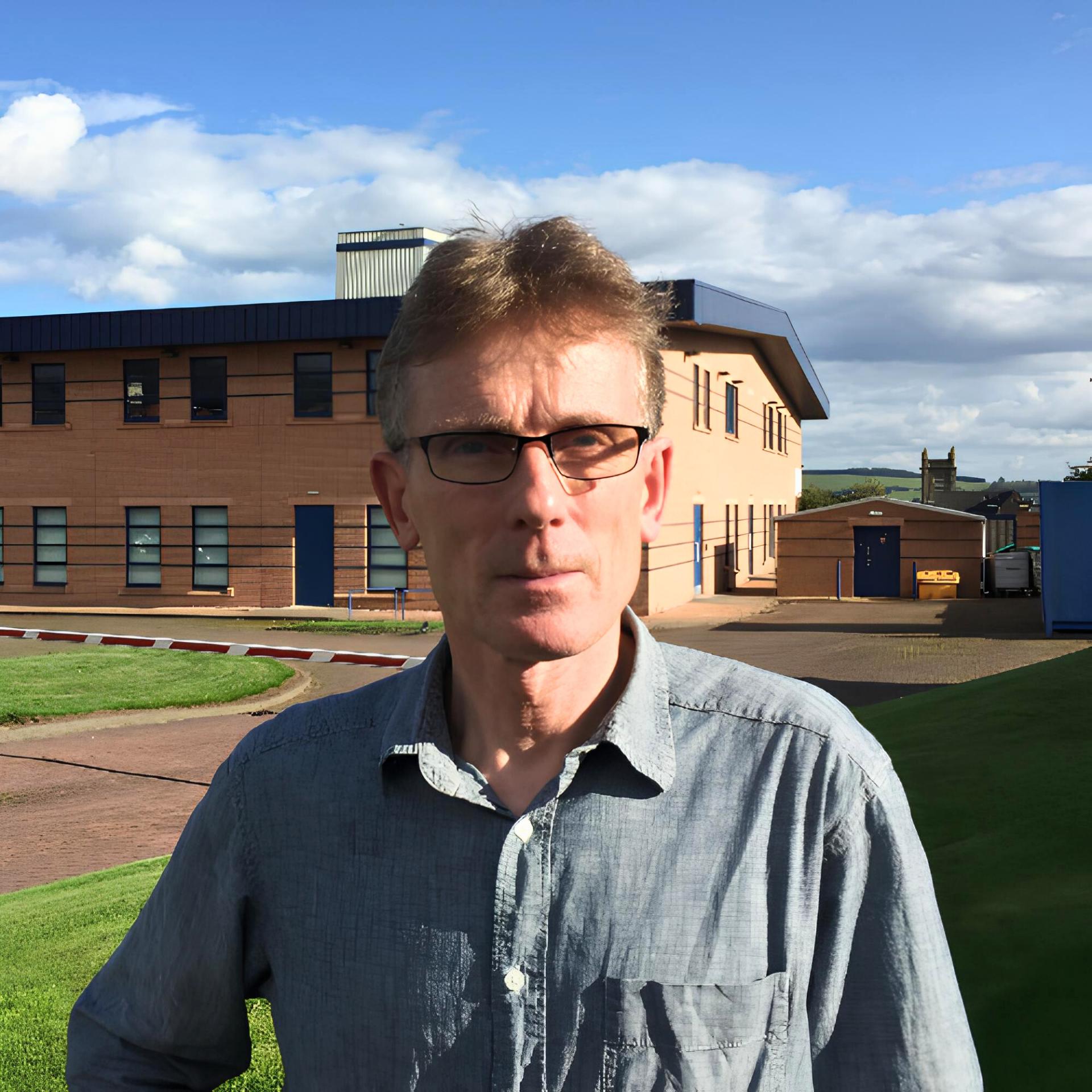 Man with glasses in a gray shirt stands outside near a building with a blue roof on a sunny day.