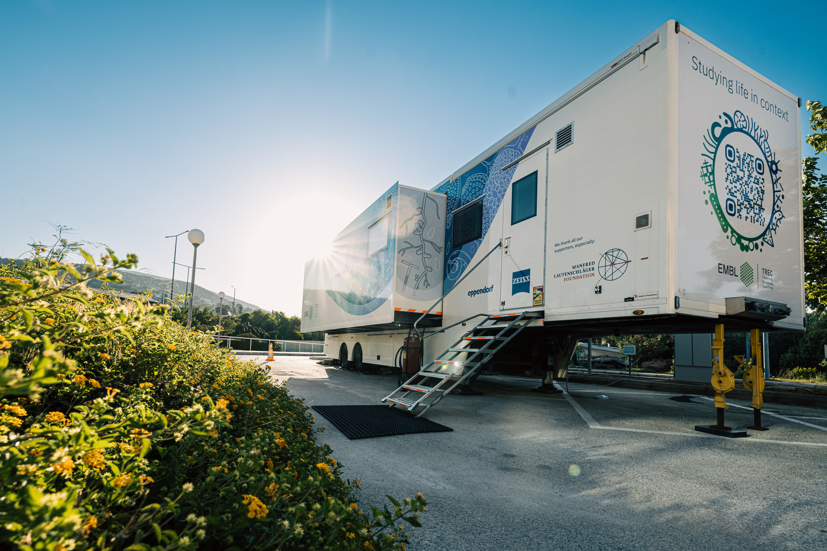 Mobile laboratory trailer parked on a sunny day, surrounded by plants, with a visible QR code and scientific graphics on its side.