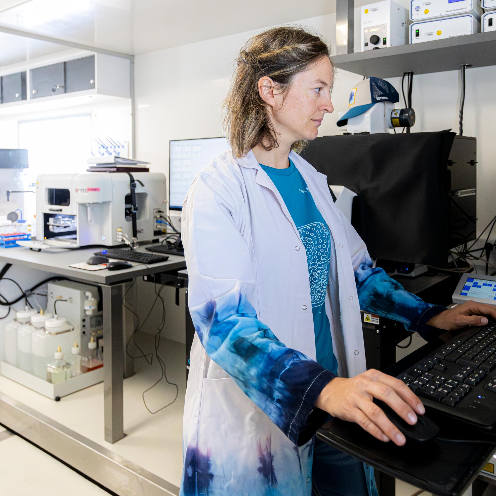 A woman in a white and blue lab coat stands at a PC and looks at the screen. Laboratory instruments can be seen in the background.
