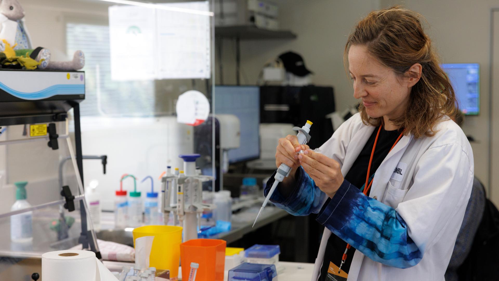 A scientist wearing a lab coat uses a pipette in a laboratory filled with equipment and supplies.