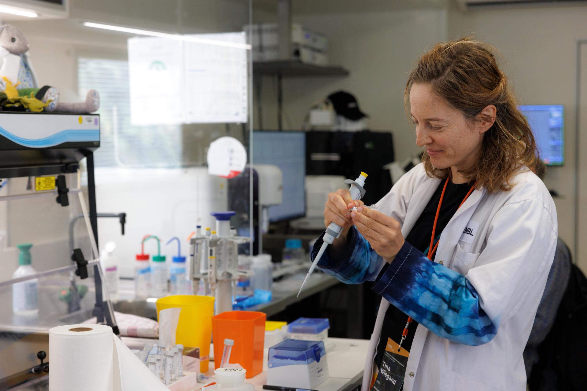 A scientist wearing a lab coat uses a pipette in a laboratory filled with equipment and supplies.