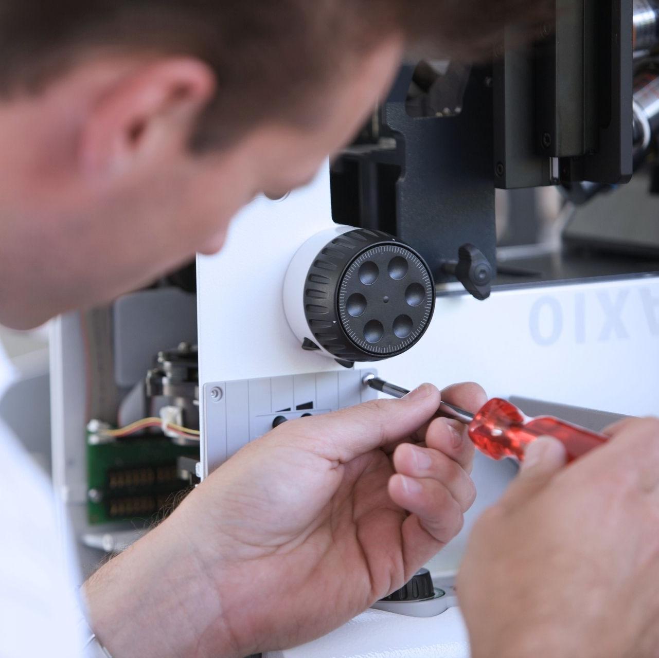 A technician using a red-handled screwdriver to make precise adjustments on a white laboratory instrument. The background shows an organized setup with visible machine components.