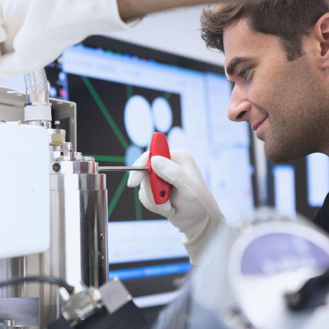 A professional wearing white gloves using a red-handled tool to adjust components on a metallic device. A computer screen displaying analysis data is visible in the background.